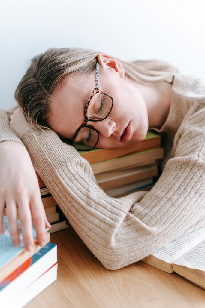 A woman in a sweater rests with books, wearing glasses. Ideal stock for themes of education and exhaustion.