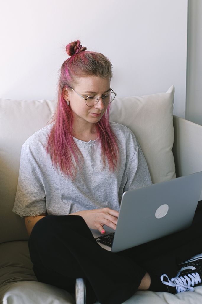 A young woman with pink hair uses her laptop while sitting on a couch, indoors.